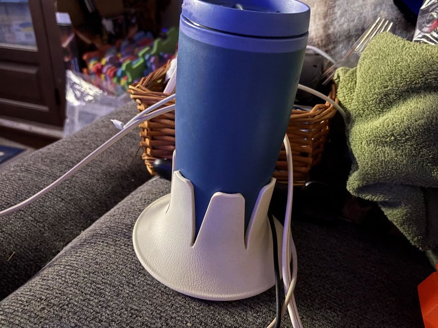 A blue travel tumbler in a cream drink base perched on the upholstered arm of a grey couch, charging cables and a folded green towel around it, dim room at night